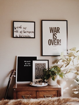 A cozy and artistic interior setting includes a wooden dresser adorned with framed art prints and a potted plant. The art on the wall features motivational and inspirational text, with phrases encouraging introspection and positivity. A guitar leans against the side, and the soft lighting gives the space a warm and inviting atmosphere.