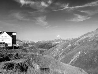 A black and white photograph features a house situated on a hillside with a backdrop of expansive mountain ranges. The sky is mostly clear with a few scattered clouds, contributing to an expansive landscape view. The house, positioned on the left side, has multiple windows and a small balcony area, providing a sense of isolation yet tranquility.