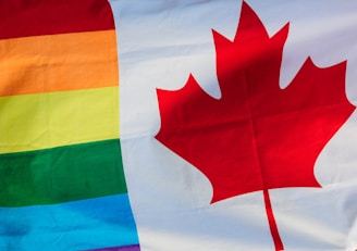 A composite flag featuring the rainbow stripes of the LGBTQ+ pride flag on the left. On the right, a large red maple leaf on a white background is prominently displayed, representing Canada.
