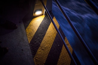 A diagonal section of a yellow and black striped warning surface illuminated by a light. The surrounding area is dark, with visible metal railings and a blurred movement of water beside it.