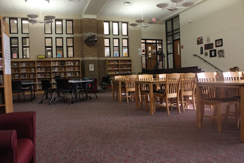A library reading area with bookshelves lining the wall, filled with books. Several tables with wooden chairs are neatly arranged around the room. A few black chairs are grouped around a small round table. The space features a high ceiling with unique light fixtures and large windows allowing natural light to flood in, creating a bright and inviting environment. A set of stairs with black railings leads up to a platform area, and several framed pictures are hung on the wall.
