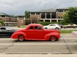 A vibrant red vintage car in motion passes by on a city street, creating a sense of action and speed. In the background, a modern office building with a Merrill Lynch sign is visible, surrounded by greenery. Several other vehicles, including a black car and a white car, are part of the traffic scene.