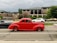A vibrant red vintage car in motion passes by on a city street, creating a sense of action and speed. In the background, a modern office building with a Merrill Lynch sign is visible, surrounded by greenery. Several other vehicles, including a black car and a white car, are part of the traffic scene.