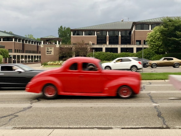 A vibrant red vintage car in motion passes by on a city street, creating a sense of action and speed. In the background, a modern office building with a Merrill Lynch sign is visible, surrounded by greenery. Several other vehicles, including a black car and a white car, are part of the traffic scene.