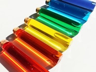 A collection of seven transparent glass test tubes filled with liquids in vibrant, distinct colors, ranging from red, orange, yellow, green, to blue. Each tube is sealed with a cork and is positioned diagonally on a clean, white surface. The arrangement creates a visually appealing gradient effect as the colors transition smoothly from warm to cool hues, casting soft shadows below.