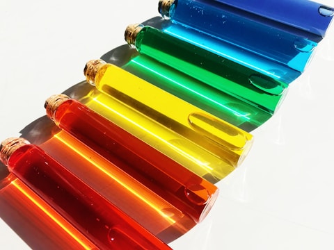 A collection of seven transparent glass test tubes filled with liquids in vibrant, distinct colors, ranging from red, orange, yellow, green, to blue. Each tube is sealed with a cork and is positioned diagonally on a clean, white surface. The arrangement creates a visually appealing gradient effect as the colors transition smoothly from warm to cool hues, casting soft shadows below.