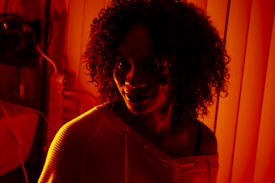 A person with curly hair stands in warm, red-toned lighting, creating a dramatic shadow effect on the face. The background consists of vertical blinds casting stripes of light and shadow across the scene.