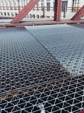 A close-up view of a metal grating walkway on a bridge. The grating has a patterned design and stretches across the bridge, with visible beams underneath. In the background, there are red metal railings and structures, as well as a building with windows.