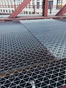 A close-up view of a metal grating walkway on a bridge. The grating has a patterned design and stretches across the bridge, with visible beams underneath. In the background, there are red metal railings and structures, as well as a building with windows.