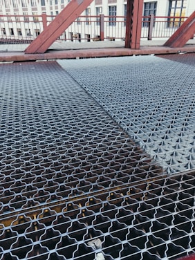 A close-up view of a metal grating walkway on a bridge. The grating has a patterned design and stretches across the bridge, with visible beams underneath. In the background, there are red metal railings and structures, as well as a building with windows.