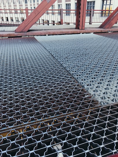 A close-up view of a metal grating walkway on a bridge. The grating has a patterned design and stretches across the bridge, with visible beams underneath. In the background, there are red metal railings and structures, as well as a building with windows.