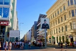 A bustling street scene features several people walking and gathering near a historical checkpoint. The architecture includes modern and classical styles, with notable buildings like a KFC and McDonald's. A prominent sign displays a military figure in uniform, while signs and advertisements adorn the surrounding buildings.