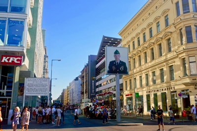 A bustling street scene features several people walking and gathering near a historical checkpoint. The architecture includes modern and classical styles, with notable buildings like a KFC and McDonald's. A prominent sign displays a military figure in uniform, while signs and advertisements adorn the surrounding buildings.