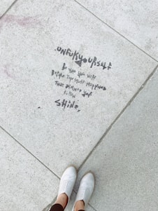 Text is written on a concrete sidewalk, saying 'unf***yourself. Be who you were before the stuff happened that dimmed your f*****g shine.' A pair of white shoes is visible at the bottom of the image, with someone standing on the pavement.