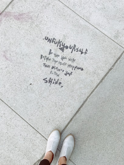 Text is written on a concrete sidewalk, saying 'unf***yourself. Be who you were before the stuff happened that dimmed your f*****g shine.' A pair of white shoes is visible at the bottom of the image, with someone standing on the pavement.