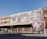 A multi-story industrial building with a weathered facade featuring a black-and-white mural. The building has numerous large, rectangular windows with a grid pattern. On the right side, there is a colorful mural with vibrant graphics. A chain-link fence surrounds the perimeter, and a few trees and bike racks are visible on the sidewalk in front.