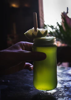 A hand holds a glass jar filled with a vibrant green liquid. The jar is topped with a slice of lime, fashioned into a decorative flower shape, secured with a wooden stick. The setting appears dimly lit, with soft focus greenery in the background, creating an artistic and moody ambiance.