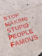 Red stencil graffiti text on a concrete sidewalk conveys a message against promoting individuals without merit. The letters are bold and slightly weathered.
