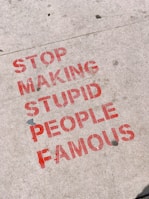 Red stencil graffiti text on a concrete sidewalk conveys a message against promoting individuals without merit. The letters are bold and slightly weathered.
