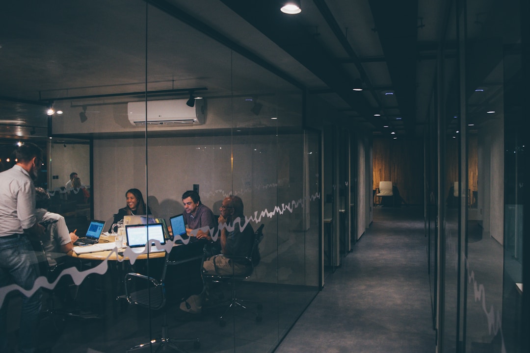 A team meeting in a conference room with glass windows, with employees on laptops discussing proposal management.