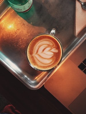 A cup of coffee with intricate latte art on a metallic tray, next to a green bottle and the edge of a laptop. The warm lighting creates a cozy atmosphere.