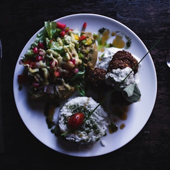 A plate featuring a colorful arrangement of various foods, including a portion of white rice garnished with a cherry tomato, falafel drizzled with a white sauce, and a fresh salad topped with diced vegetables and a creamy dressing. The foods are arranged aesthetically on a round white plate placed on a dark surface.