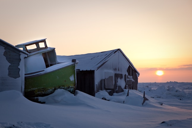 An old, snow-covered boat leans against a weathered wooden shed, both blanketed with snow in a wintry landscape. The sun is setting on the horizon, casting a warm glow over the icy scene.