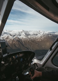 Aerial view of snow-capped mountains seen through the cockpit of an aircraft. The cockpit includes various control instruments and a smartphone mounted on a holder. Part of the pilot's hand gripping a control stick is visible.