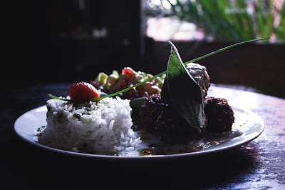 A white plate holds a serving of white rice with herbs, accompanied by several deep-fried balls, possibly falafel, adorned with a green leaf. A cherry tomato and mixed vegetables or salad add color to the dish. The background is dark, with glimpses of green foliage.