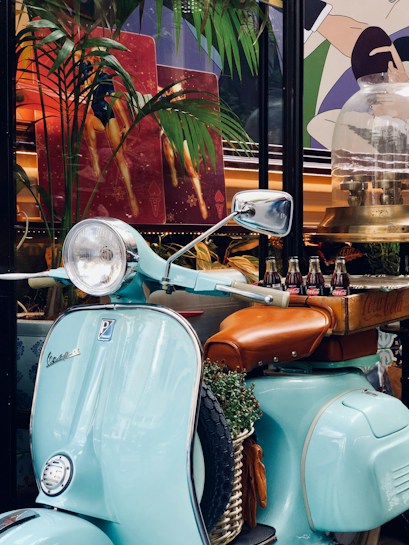 A vintage light blue scooter with a brown leather seat is parked, adorned with greenery in a basket at the front. Behind it is a box with glass bottles placed on the seat. The setting is tropical and lively with palm leaves and vibrant artwork in the background, featuring colorful geometric shapes and figures.