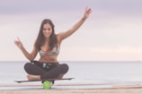 A woman is balancing on a board supported by a cylindrical roller, sitting cross-legged with her arms raised. The setting is a beach with calm water in the background under a cloudy sky. She appears to be focused and confident.