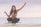 A woman is balancing on a board supported by a cylindrical roller, sitting cross-legged with her arms raised. The setting is a beach with calm water in the background under a cloudy sky. She appears to be focused and confident.