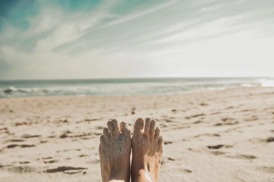 Two feet lie on sandy beach, toes pointed upwards, with sandals visible in foreground