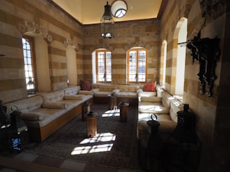 A traditional room with beige brick walls and high ceilings features arched windows that let natural light stream in. The seating area includes beige cushioned benches along the walls and decorative pillows. Intricately carved woodwork and lantern-style lamps add to the decor. The floor is covered with a patterned rug.