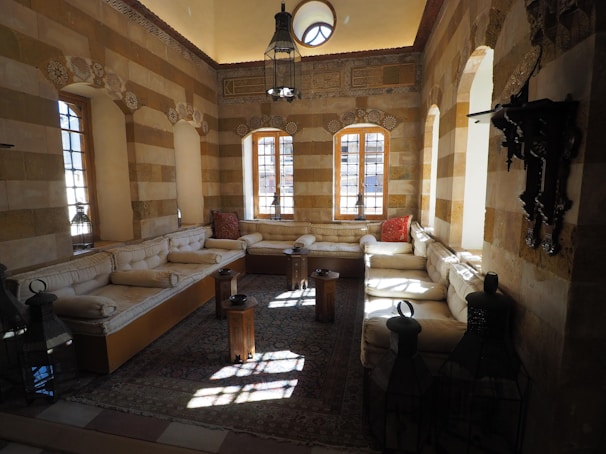 A traditional room with beige brick walls and high ceilings features arched windows that let natural light stream in. The seating area includes beige cushioned benches along the walls and decorative pillows. Intricately carved woodwork and lantern-style lamps add to the decor. The floor is covered with a patterned rug.