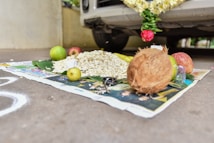 An arrangement of ritual items placed on a newspaper in front of a car. The setup includes a green banana leaf with a mound of puffed rice, a whole coconut, several fruits such as apples and limes, a bunch of small keys, and some flowers attached to the car. The scene suggests a ceremonial offering or blessing for the vehicle.