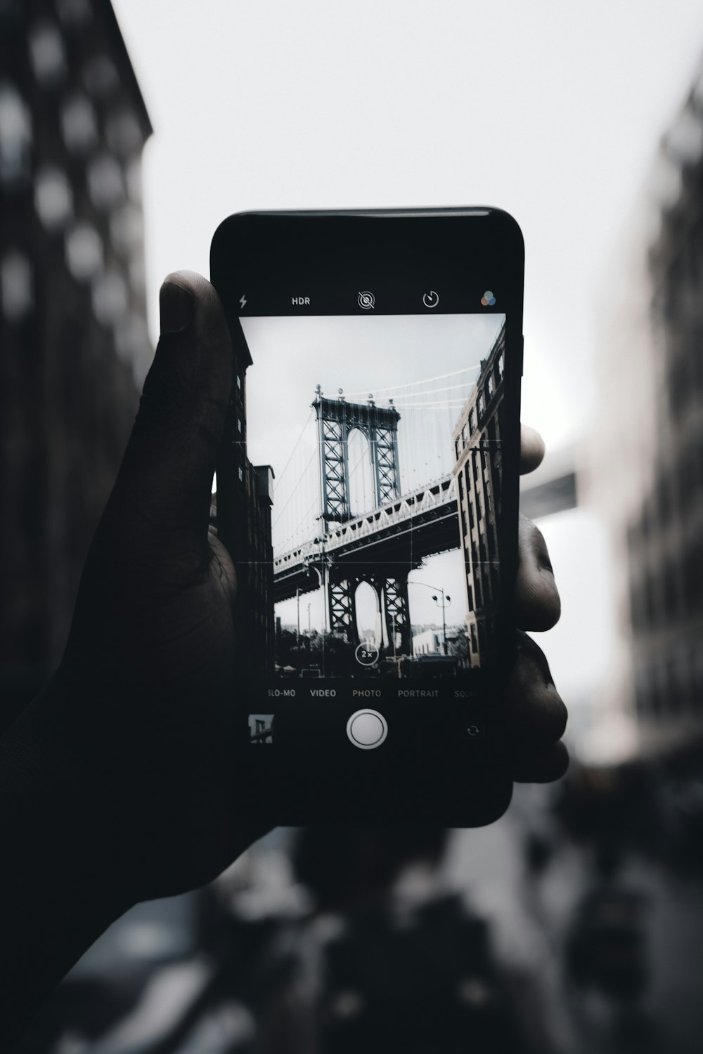 grayscale photography of person holding iPhone capturing bridge