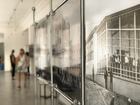 An art gallery with blurred figures of people in the background and a sharp focus on framed black and white photographs of historical scenes on the right. The space is filled with soft, natural light.