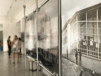 An art gallery with blurred figures of people in the background and a sharp focus on framed black and white photographs of historical scenes on the right. The space is filled with soft, natural light.