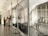 An art gallery with blurred figures of people in the background and a sharp focus on framed black and white photographs of historical scenes on the right. The space is filled with soft, natural light.