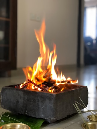 A small fire burns brightly in a square, stone container on a smooth floor. The flames are vivid, casting a warm glow. Several sticks are visible within the flames, and a brass bowl and green leaf are nearby, suggesting a ritual or ceremonial setting.