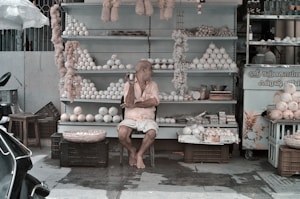 A street vendor sits in front of a stall filled with a variety of items, including white spherical objects and woven baskets. The vendor appears relaxed, sitting on a stool with legs crossed, while wearing casual clothing. There are shelves in the background and some items, like garlands, hang from above.