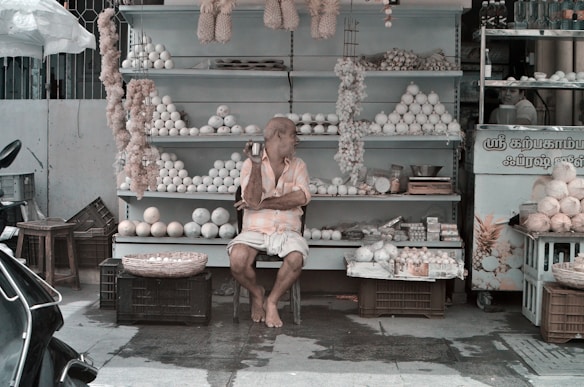 A street vendor sits in front of a stall filled with a variety of items, including white spherical objects and woven baskets. The vendor appears relaxed, sitting on a stool with legs crossed, while wearing casual clothing. There are shelves in the background and some items, like garlands, hang from above.