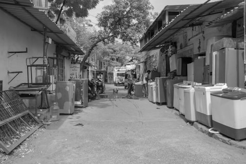 A narrow street lined with various household appliances such as washing machines and refrigerators. The appliances are placed on either side of the street, outside small shops with corrugated roofs. A few people can be seen engaging in different activities, including a person on a motorbike. A dog stands in the middle of the street. Trees and branches stretch overhead, providing some shade.