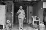 An elderly man stands in a doorway while looking down at a smartphone. He holds a small bag in his other hand. The setting appears to be a modest and aged interior space with visible kitchen appliances and gas cylinders. There is a plastic chair to the right, and the overall environment seems utilitarian with worn surfaces.