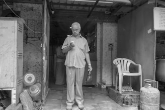 An elderly man stands in a doorway while looking down at a smartphone. He holds a small bag in his other hand. The setting appears to be a modest and aged interior space with visible kitchen appliances and gas cylinders. There is a plastic chair to the right, and the overall environment seems utilitarian with worn surfaces.