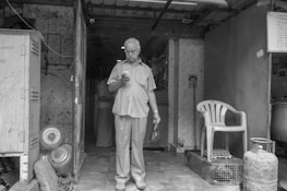 An elderly man stands in a doorway while looking down at a smartphone. He holds a small bag in his other hand. The setting appears to be a modest and aged interior space with visible kitchen appliances and gas cylinders. There is a plastic chair to the right, and the overall environment seems utilitarian with worn surfaces.