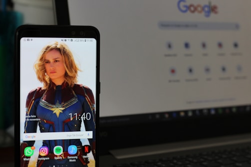 A smartphone displays a wallpaper featuring a blonde woman in a blue and red superhero costume. The device's screen shows various app icons such as WhatsApp, Instagram, Spotify, and Netflix. In the background, a laptop screen displays a Google search page.