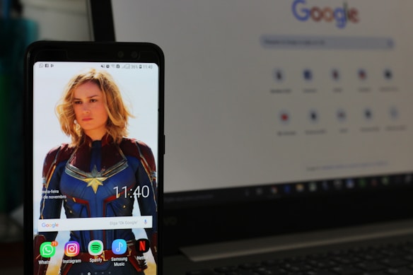 A smartphone displays a wallpaper featuring a blonde woman in a blue and red superhero costume. The device's screen shows various app icons such as WhatsApp, Instagram, Spotify, and Netflix. In the background, a laptop screen displays a Google search page.