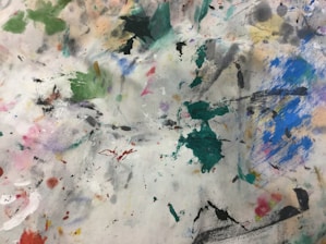 A chaotic and colorful array of paint splatters and brush strokes covers a surface. The vibrant mix includes shades of green, blue, pink, and black, creating an abstract pattern. The texture and overlapping colors suggest a well-used artist's palette or workspace.